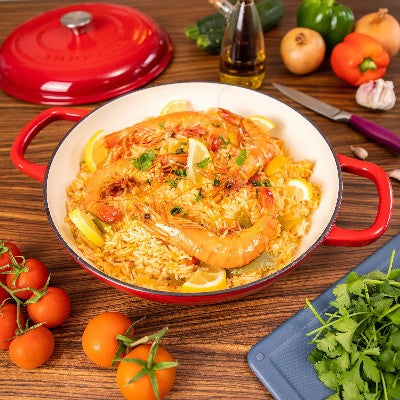 poele_en_fonte_emaillee rouge_avec_deux_anses_ sur table bois avec epices autour et paella dedans 